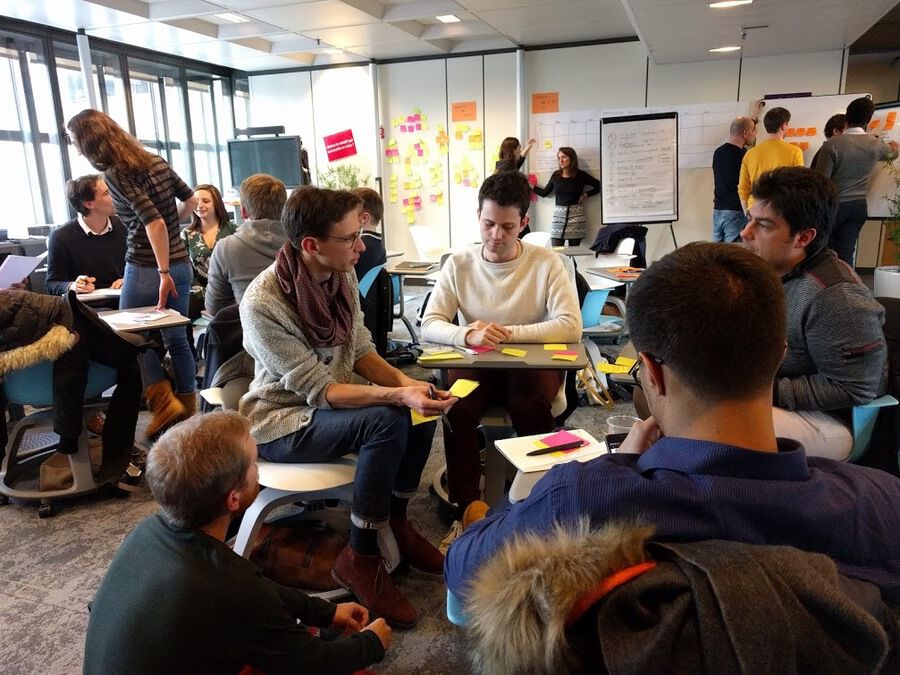 Photo représentant une dizaine de personnes, qui font des activités en petits groupes : discuter, écrire sur des posts-its, écrire sur un tableau.