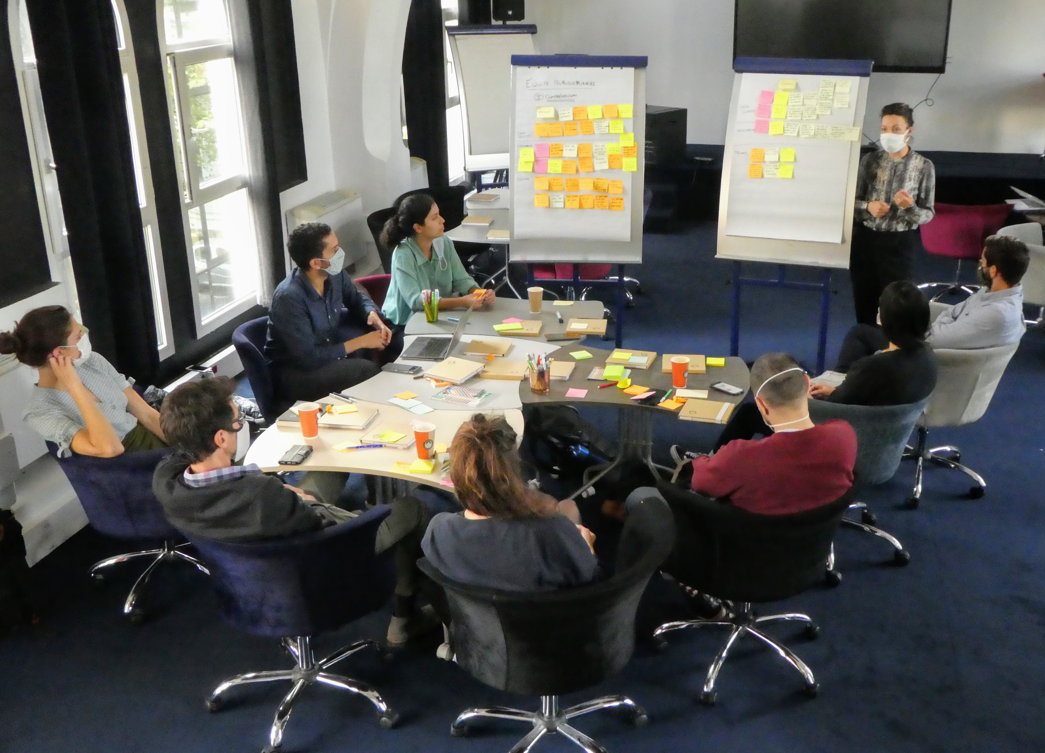 Huit personnes sont assises autour d’une table. Une personne présente. Les huit personnes font face à deux tableaux blancs, sur lesquels ont été collés des post-its.