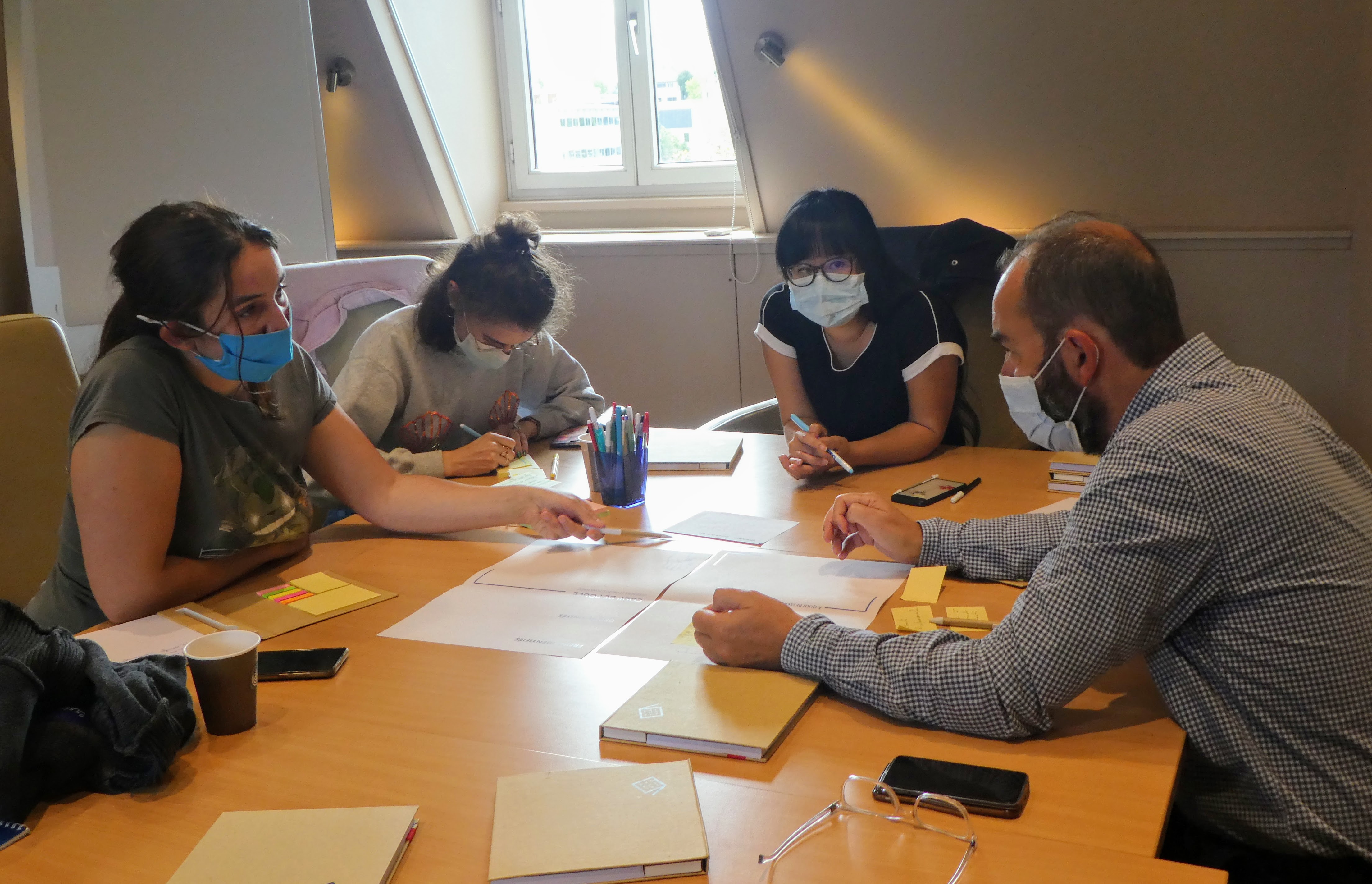 Quatre personnes sont réunies autour d’une table. Une personne écrit sur un post-it. Trois personnes sont engagés dans une discussion