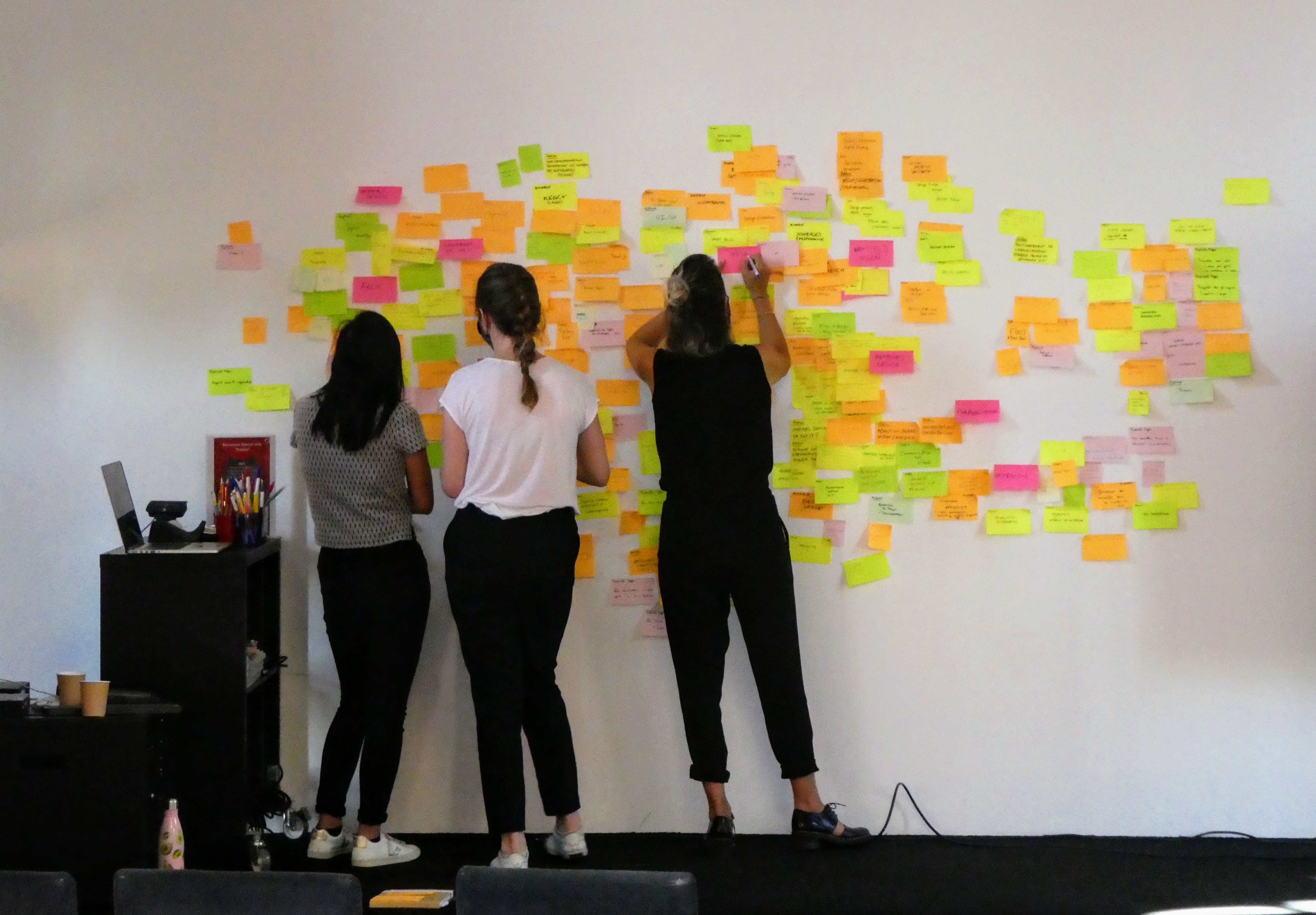 Un mur blanc est couvert de post-its de quatre couleurs différentes. Trois personnes lui font face. Une personne écrit sur un post-it rose