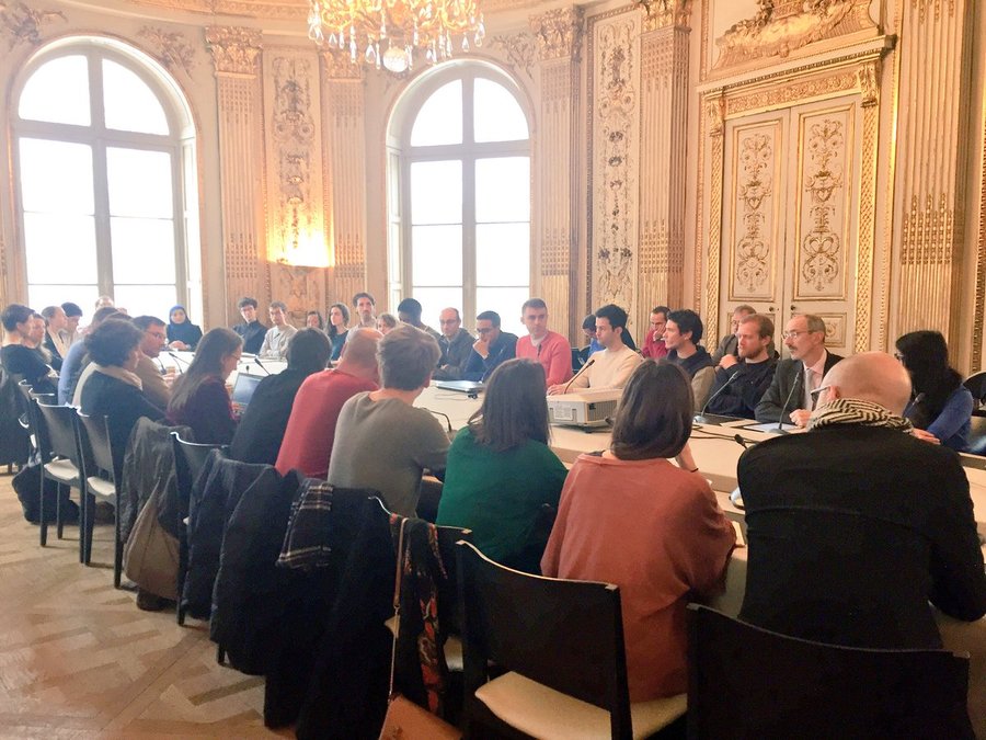 Plan d’ensemble d’une quarantaine de personnes autour d’une table, dans une salle de réunion agrémentée de dorures.