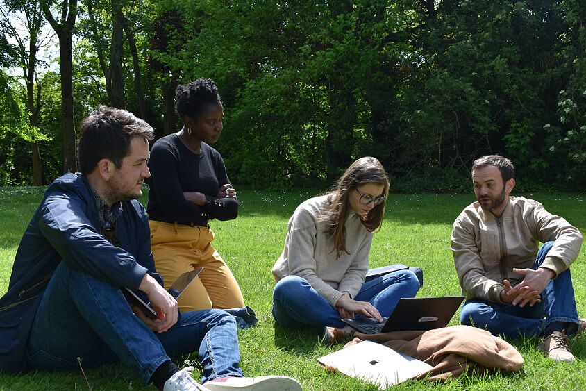 Deux hommes et deux femmes sont assis dans l’herbe avec leurs ordinateurs. Tous écoutent l’homme assis à l’extrême droite de la photo.