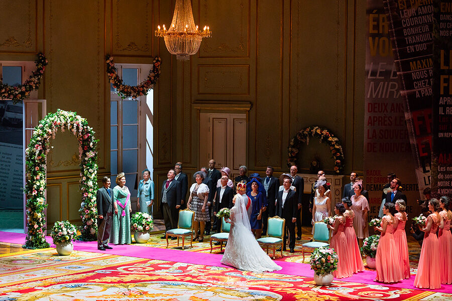 Une scène d’opéra. Au premier plan, un tapis de style Empire, rouge avec des motifs jaunes et bleus. Dessus, un couple de mariés s’avance en direction d’une arche fleurie.